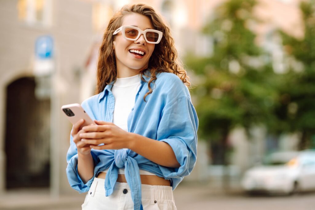 Fröhliche junge Frau mit modischer Sonnenbrille beschäftigt sich mit ihrem Smartphone und genießt das schöne Wetter auf einer belebten Stadtstraße voller Grün.